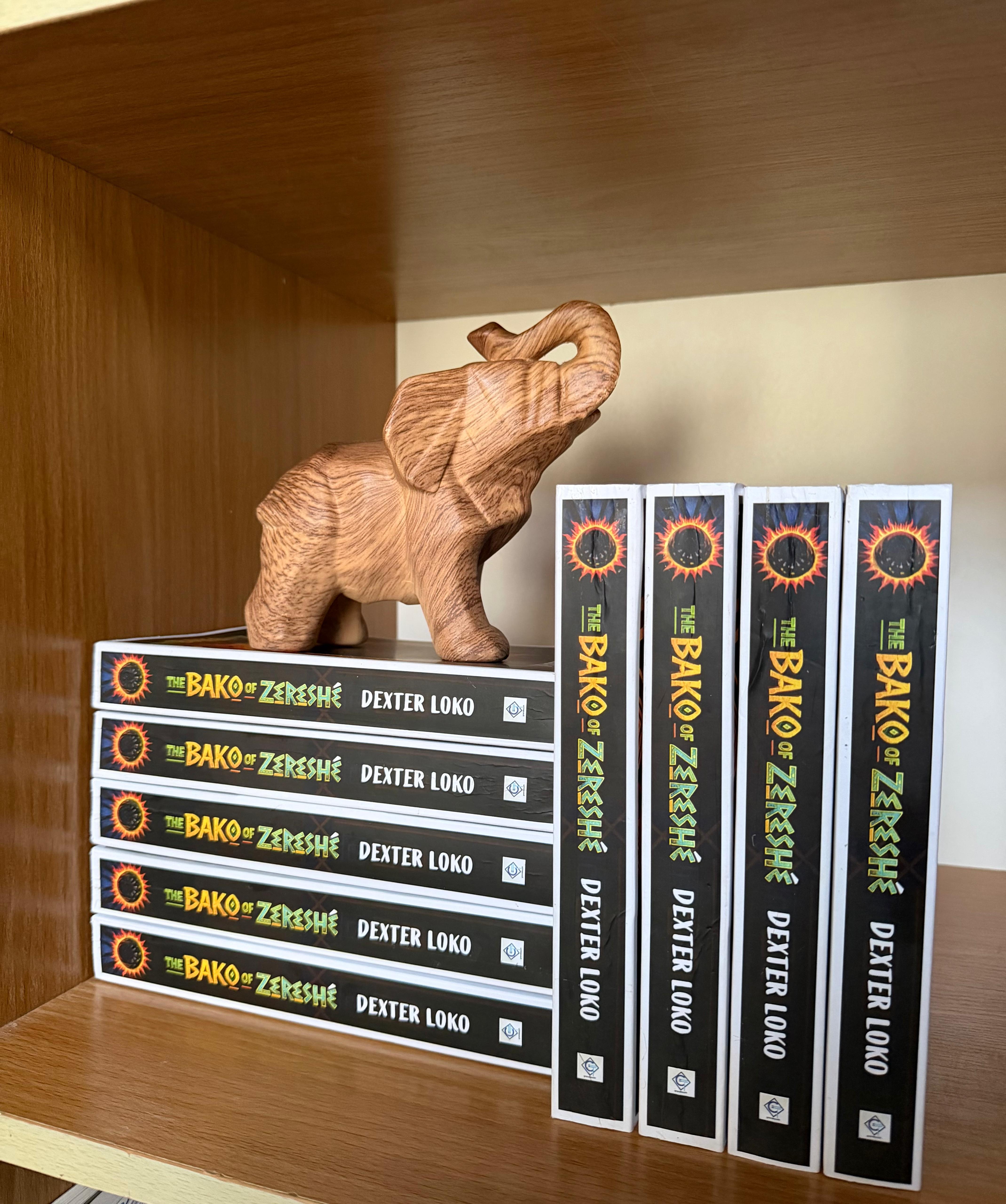 Copies of The Bako of Zereshé arranged on a wooden shelf beneath a carved elephant.